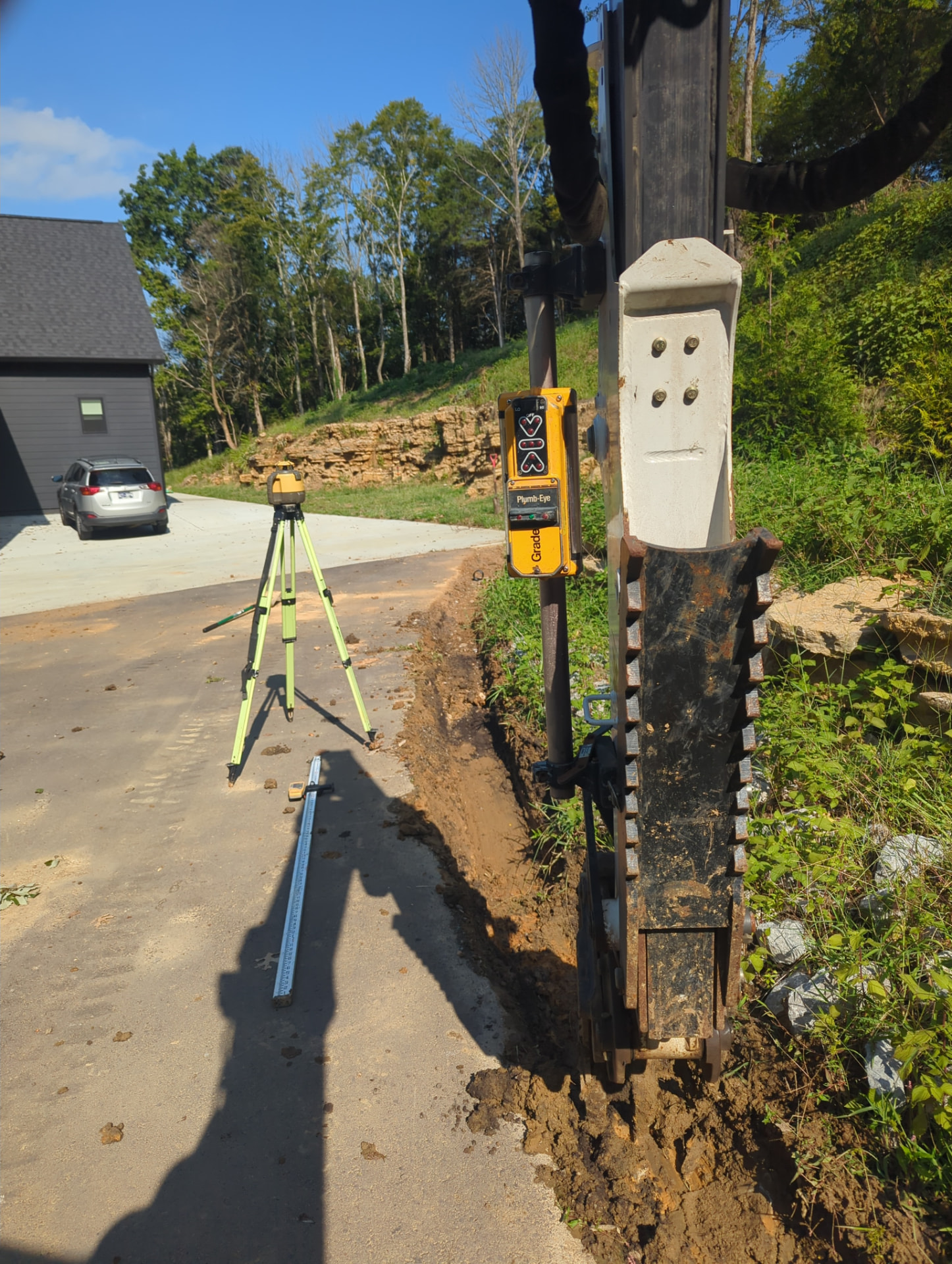 Laser level and machine-mounted grade receiver beside a driveway drainage trench