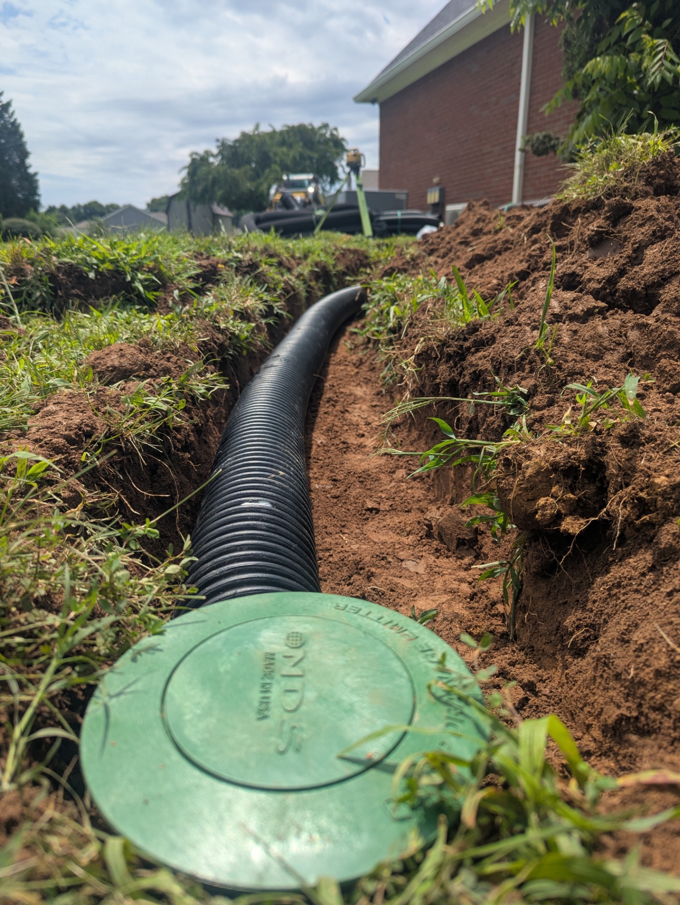 Black corrugated downspout extension pipe running through a trench to a green NDS popup emitter beside a home